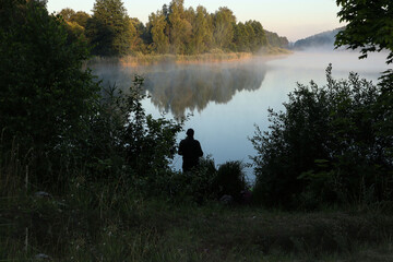 Silhouette of a fisherman against the backdrop of a foggy lake on a summer morning
