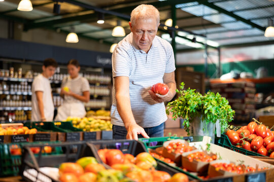Interested elderly man shopping for organic vegetables in local farm store, choosing ripe tomatoes on produce display..