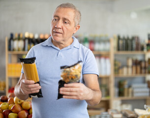 Uncertain mature male buyer choosing a package of pasta or spaghetti in grocery store