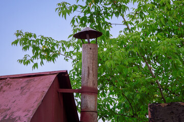 A rustic wooden chimney featuring a metal cap rises above a weathered roof, framed by vibrant green foliage under a blue sky
