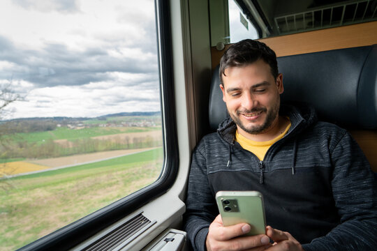 Hombre sonriente usando su celular mientras viaja en tren, sentado en la ventanilla