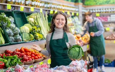 Positive young female seller holding cabbage standing by counter in vegetable market