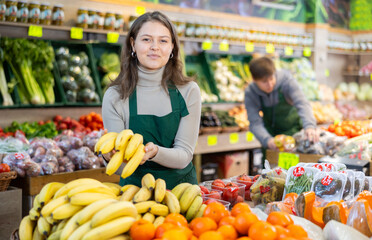 Happy young shop assistant setting bunch of bananas on a counter in big greengrocery