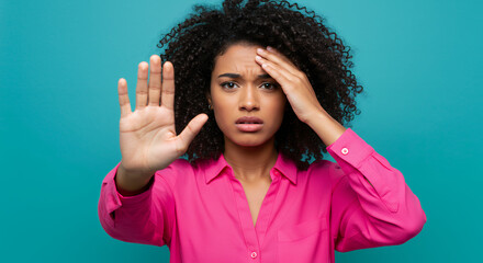 Distressed Black woman in her 20s holding hand up in a stop gesture with a concerned expression and her other hand on her forehead against teal background