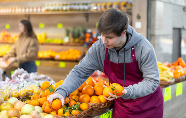 Young salesman in apron lays out tangerines on counter in vegetable shop
