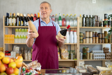 Polite mature male seller kindly offering bottled wine in cozy grocery store