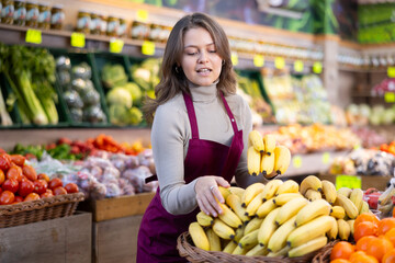 Young positive female seller in apron displaying assortment of bananas at supermarket