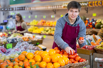 Young salesman in apron lays out tomatoes on counter in vegetable shop..