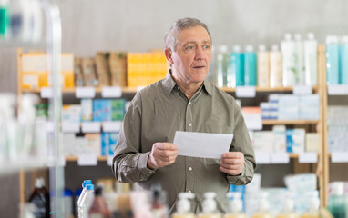 Senior man with a medical prescription stands near a counter with medicines and pills in a European...