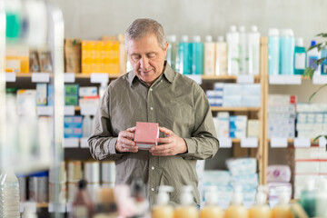 Senior man buys emollient cream at pharmacy and compare with other medicine, carefully reads...