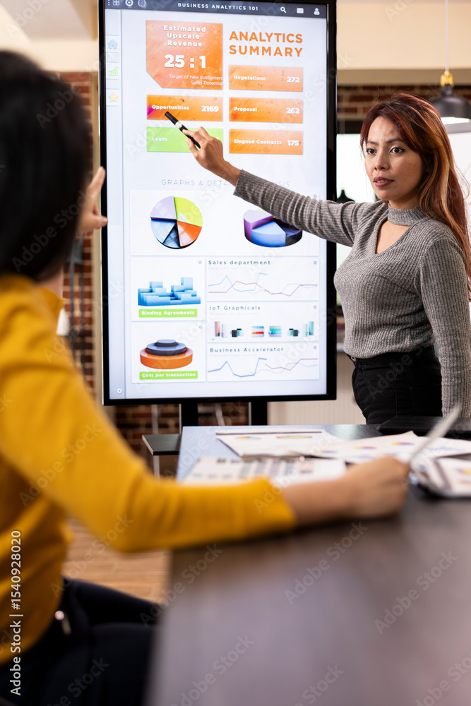 Poster female analyst presents strategies to colleague, using digital monitor to display company analytics 