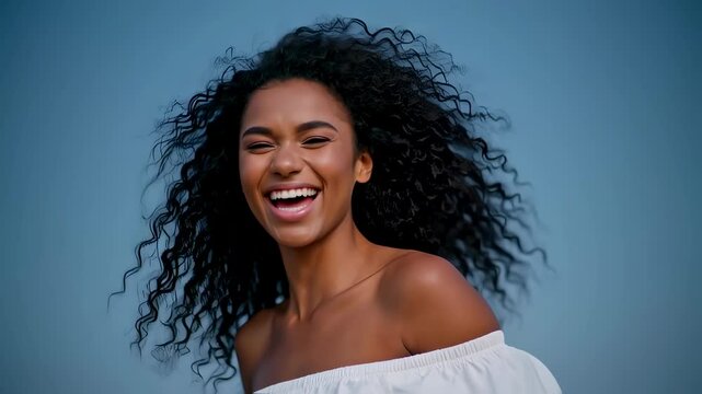  A beautiful, happy young woman with dark curly hair blowing in the wind laughs with a radiant smile. This medium shot captures her carefree spirit against a simple blue background.