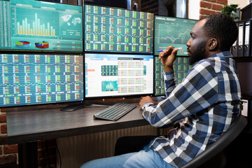 African american stock trader working late, tracking market trends on a multiple monitor...