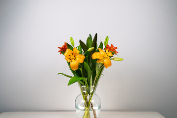 A fresh floral arrangement resting in a clear glass vessel, showcasing vibrant red and orange blooms, long green leaves, and budding flowers
