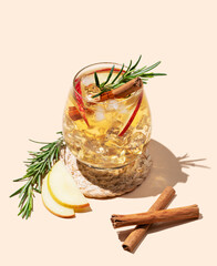 Apple cocktail or cider in a glass with ice, cinnamon and rosemary on a beige background