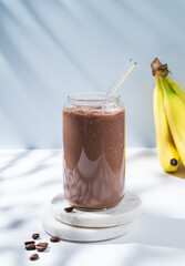 Chocolate coffee smoothie with banana in a glass on a white blue background with morning shadow.