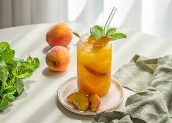 Peach drink or tea with ice, mint in a glass on a white table near window with fresh fruits, herbs