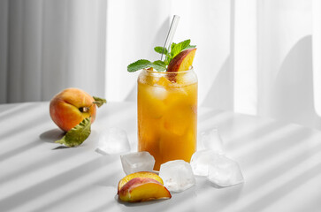 Refreshing peach cocktail, lemonade or iced tea on a white table near window with shadows and ice cubes.