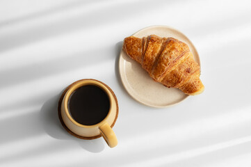 Coffee espresso in yellow cup and fresh croissant on white background with morning shadows.