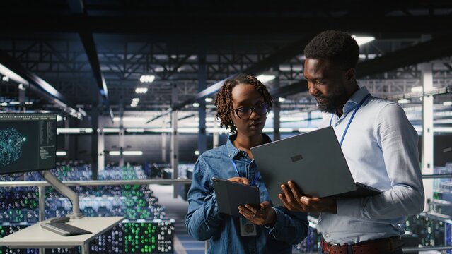 Data center system administrators using laptop to review AI LLM visualizations. African american IT specialists use device to analyze neural networks outputs, ensuring stable gear operations