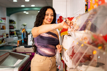 A Latina girl between 20 and 30 years old is shopping in a small business.The woman is picking up candies from some transparent boxes.The concept of buying in small retail stores.