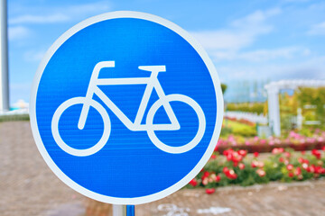 A blue and white traffic sign displaying a bicycle symbol against a backdrop of the sky and clouds. The image includes a bicycle wheel, tire, frame, and a plant in the background