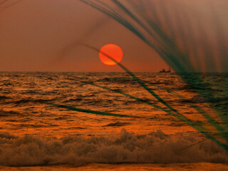 A breathtaking sunset or sunrise with a calm sea and big red sun setting down the horizon. A tranquil atmosphere on a tropical coast, shore. The silhouette of a ship is visible in the distance.