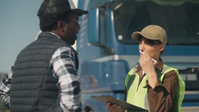 Confident African American man attentively listening to focused female in cap holding clipboard. Explaining schedule or clarifying shipping details while standing beside large blue transport truck.