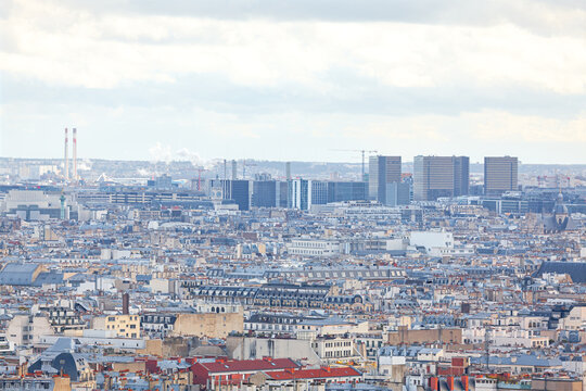 View of Paris with its iconic rooftops stretching toward modern skyscrapers in distance under a cloudy sky. City blend of historic architecture and contemporary buildings, including distant chimneys