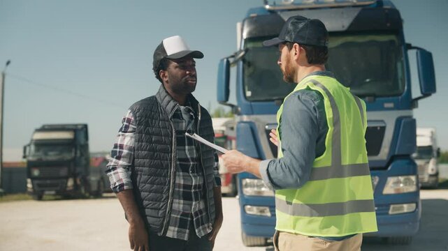 Two serious men standing near truck discussing delivery task. One holding clipboard and writing notes while second listening attentively. Reviewing shipment status or planning next logistic steps.