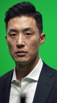 An Asian man standing without saying a word, very well dressed and with his hair done, just staring at the camera, against a green screen background.