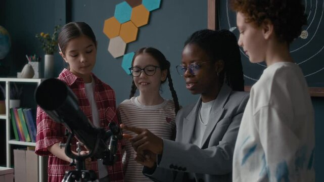 Waist up of Black female teacher in grey suit helping group of curious students operating telescope during hands-on astronomy class