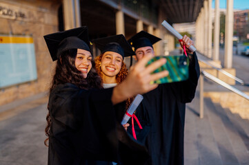 Group of cheerful multi ethnic graduates in academic gowns taking selfie with smartphone after...