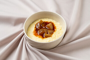 A creamy dessert topped with caramelized pear pieces sits elegantly in a simple bowl, resting on draped soft fabric in muted tones. This inviting dish evokes warmth and comfort