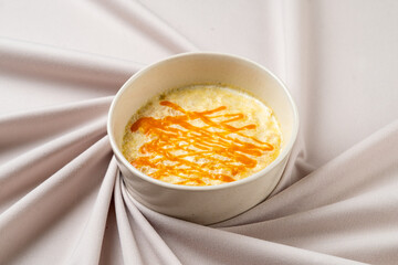 A delightful dessert placed in a pristine bowl showcases a creamy texture topped with a radiant orange drizzle, beautifully complemented by a soft fabric background