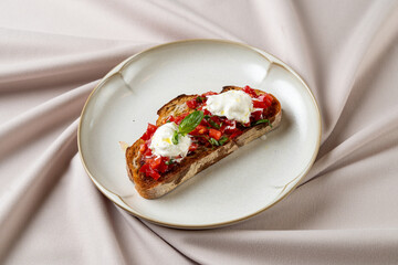 A beautifully arranged slice of grilled bread features vibrant diced tomatoes and dollops of rich cheese. This appetizing dish rests on a stylish plate against a soft, textured backdrop