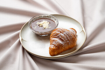 A freshly baked croissant rests on a smooth, elegant plate beside a delicate bowl filled with creamy chocolate dip
