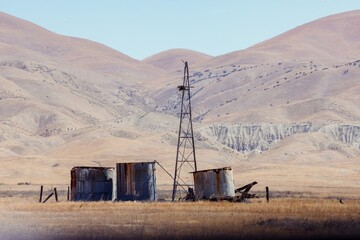 A weathered windmill with a bird's nest stands tall amidst rusted water tanks in a dry, grassy...