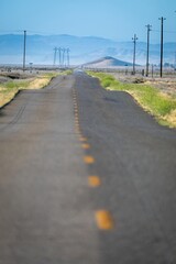 A long, straight road stretches through a rural landscape in Carrizo Plain National Monument, California, USA, marked with yellow lines. Power lines and mountains are visible in the distance.