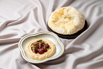 A delicious dish of creamy hummus topped with vibrant vegetables sits elegantly beside fluffy pita bread. The arrangement showcases culinary artistry in a minimalist setting