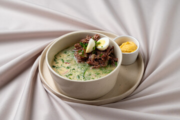 A steaming bowl of rich, flavorful soup brimming with tender meat, fresh herbs, and colorful vegetables sits on a soft fabric
