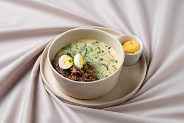 A bowl brimming with creamy soup features tender meat, sliced boiled eggs, and vibrant green herbs. A small dish of mustard rests nearby, adding a tangy touch to this inviting dish