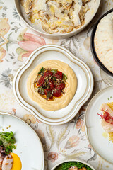 A delicious array of Middle Eastern dishes is beautifully arranged, showcasing creamy hummus topped with fresh herbs and tomatoes, offering a feast for the senses against a lovely floral backdrop