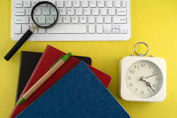 Top view white keyboard, desk clock, colorful stacked books, and a magnifying glass on a vibrant...