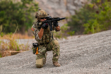 Special forces soldier uses a radio during a tactical operation while red flare smoke spreads across the rocky ground. Team communicates under high-alert battlefield conditions.