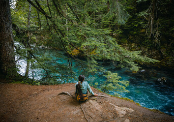 Person in Green Jacket with Yellow Backpack Sitting by Forest River