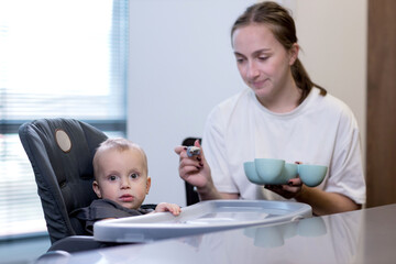 A mother is feeding her baby who is seated in a high chair using a spoon