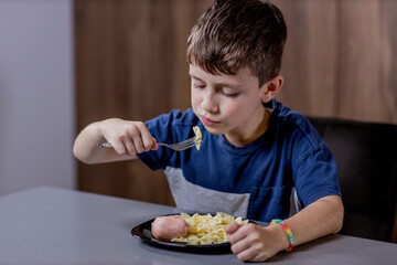 Little boy is happily eating pasta farfalle with sausage in the kitchen