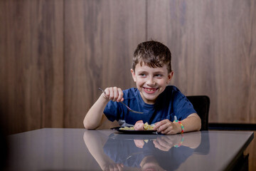 Little boy is happily eating pasta farfalle with sausage in the kitchen