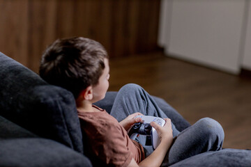 Boy is sitting on sofa at home and playing video games with his joypad.
